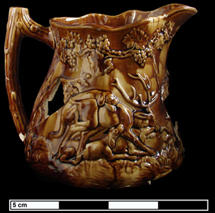 Side view of a molded yellow and brown pitcher with a scene of dogs hunting a large deer.